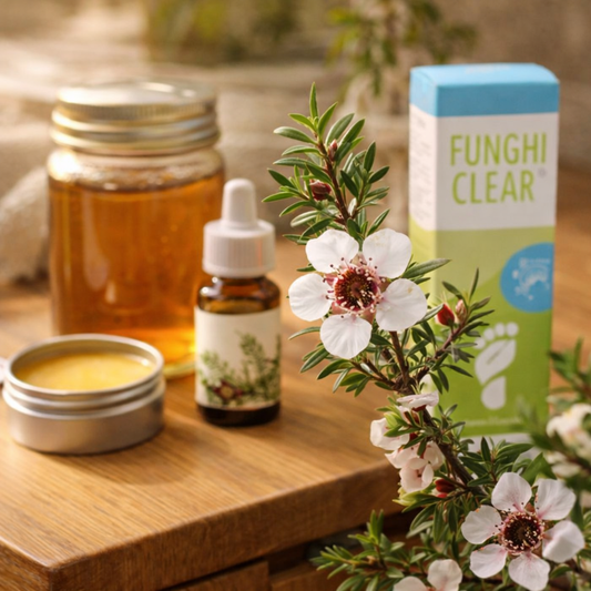 Manuka bush stems and leafy sprigs arranged on a wooden vanity beside a jar of manuka honey, a small bottle labeled manuka oil, a tin of manuka balm, and a FunghiClear box in warm natural light.